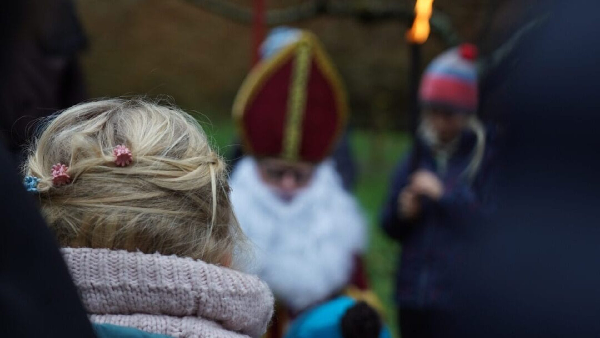 Das Bild zeigt Kinder, die Fakeln in ihren Händen halten und um Sankt Nikolaus geschart sind. Die Szenerie findet in abendlicher Dämmerung statt.