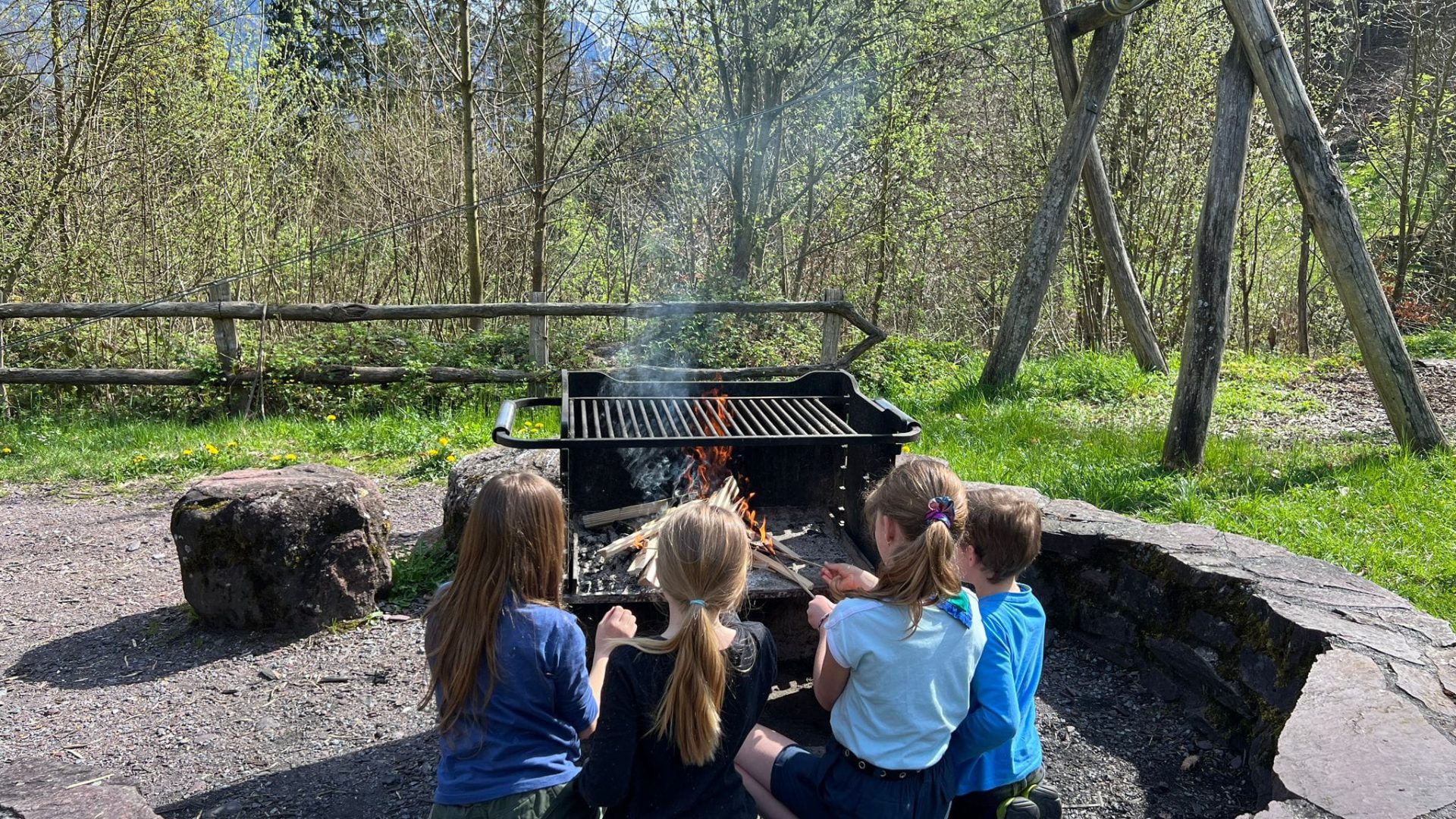 Das Bild zeigt ein paar kleine Pfadfinder, die gemeinsam Würste über einer Feuerstelle im Wald braten.