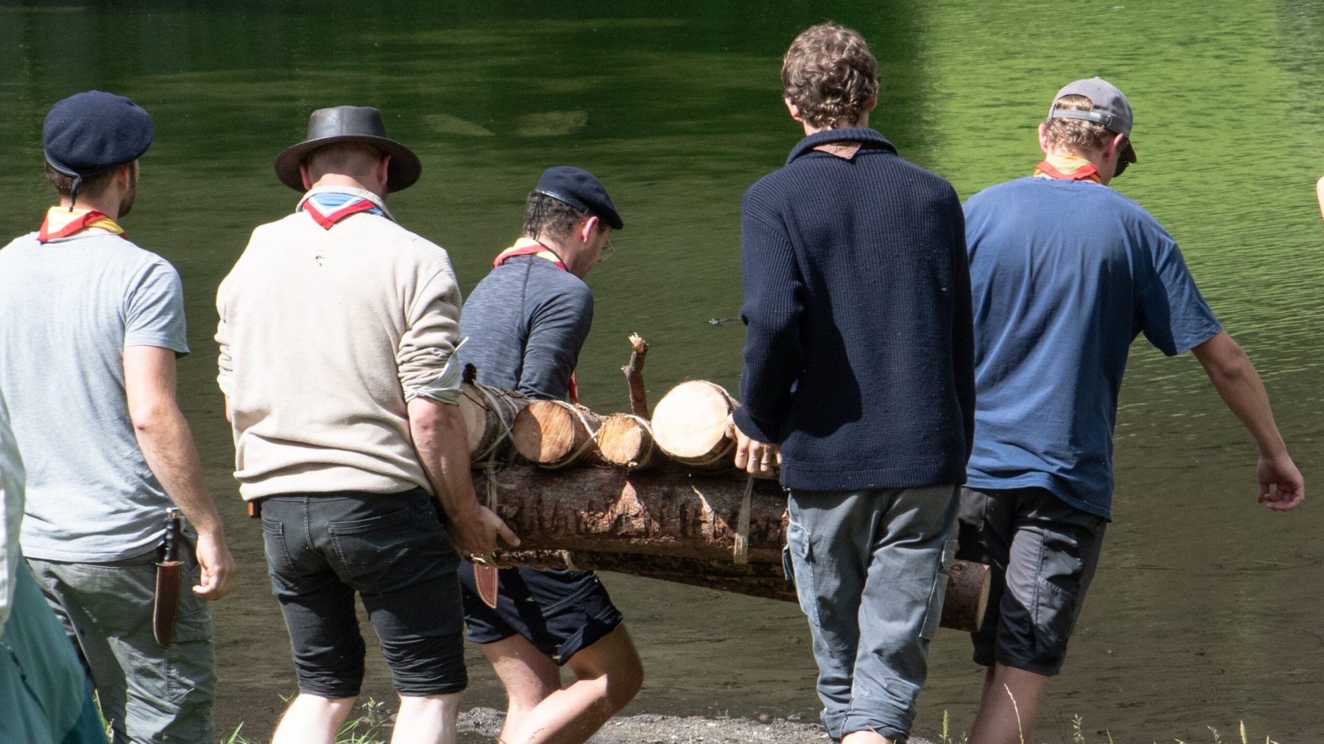 Titelbild für den Beitrag «Survivallager» (Juli 2024) Das Bild zeigt eine Gruppe Pfadfinder, die gemeinsam ein selbstgebautes Holzfloss zum Wasser tragen.